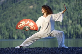 Young woman with a red hand fan practicing Taiji Pu Bu stance by the lake in the nature