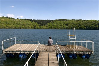 Adult woman back sitting on a pontoon at the edge of a lake surrounded by rows of trees