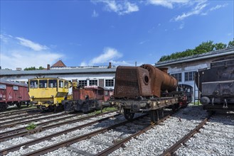 Various locomotives and a rusty hull of a steam locomotive in front of a roundhouse