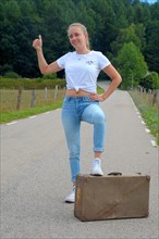 Young hitchhiking woman standing with one foot on an old vintage suitcase on a country road