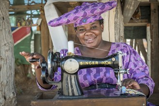 Native Herero woman with typical headgear and clothes is sitting at a sewing machine
