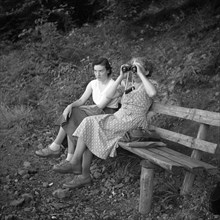 Two women are sitting on a bench and one of them is looking through binoculars