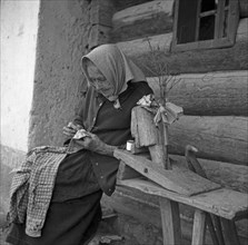 An elderly woman is sitting on a bench and knitting a shirt