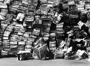 Woman reads newspaper in front of a big pile of newspapers