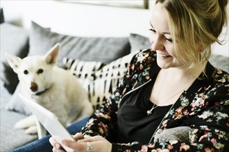 Young woman sits on the sofa with her dog and looks at a tablet computer