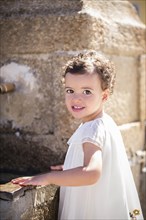 Little girl in white dress stands by a fountain