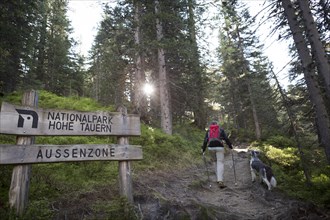 A hiker and his dog walk along a hiking trail at the entrance sign to the Hohe Tauern National Park