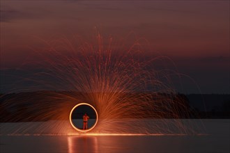 A man stands on a platform in a lake and creates a hoop of fire with glowing steel wool around him