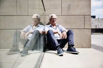 Grey-haired senior on the floor in front of a wall and reflected in a glass pane