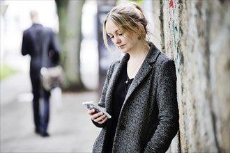 Young woman leans against a wall and looks at her smartphone