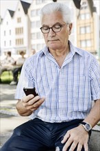 Grey-haired senior sits on a wall with his smartphone in his hand