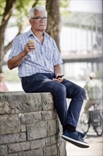 Senior sits with his Smartphone and a Coffe to go on a wall at Cologne's Rhine bank Promenade