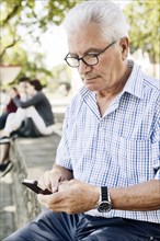 Grey-haired senior sits on a wall with his smartphone in his hand