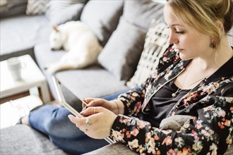Young woman sits on the sofa with her dog and looks at a tablet computer