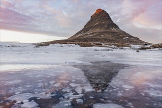 Mount Kirkjufell is reflected in the frozen lake