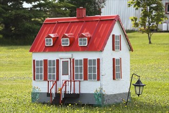 White with red trim miniature reproduction of an old Canadiana cottage style home facade in a field of yellow dandelion flowers in late spring