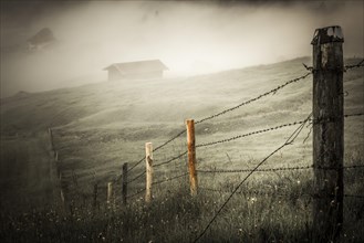 Pasture fence on mountain meadow with small cabin in the fog
