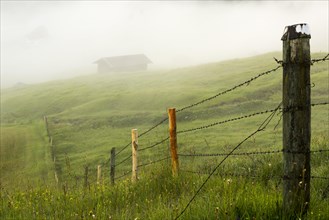 Pasture fence on mountain meadow with small cabin in the fog