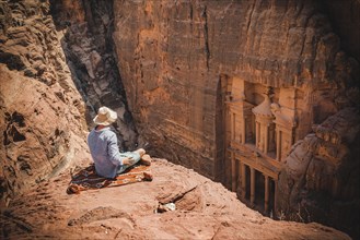 Tourist with sun hat sits on rocks and looks from above into the canyon Siq