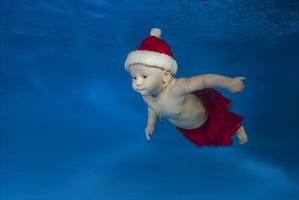 Baby in Santa's red cap swimming underwater in a pool