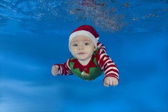 Baby boy dressed as Santa's helper swimming underwater in a pool