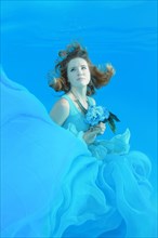 Young woman in blue dress with bouquet of flowers posing under water