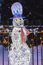 Illuminated decorative snowman attached to wrought iron fence at dusk in winter