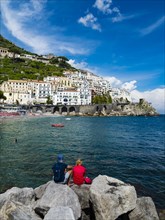 Couple sits on rock and looks at old town and beach of Amalfi
