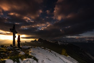 Hiker next to cross on Zafernhorn summit at sunrise with dark clouds