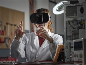 Woman with VR goggles and lab coat sitting in an electronics laboratory