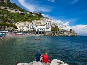 Couple sits on rock and looks at old town and beach of Amalfi