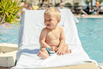 Blond toddler sitting on the sun bed by swimming pool in the resort
