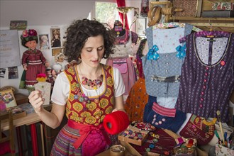 Button maker in colorful dirndl with Collier button trimmings in handicraft studio working with red yarn and utensils for Posamentenknopfe