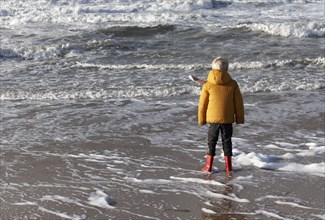 Little boy in winter clothes standing in the surf on the beach