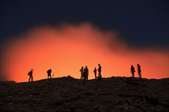 Tourists at the crater rim of the volcano Erta Ale in front of a red glowing steam cloud
