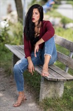 Young woman with a wine-red jacket and blue jeans sitting on a wooden bench