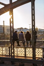 Three youths sitting on the balustrade of the Hackerbrucke bridge over the railway tracks looking into the distance