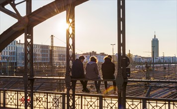 Three youths sitting on the balustrade of the Hackerbrucke bridge over the railway tracks looking into the distance