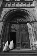 Monks with white frocks walk through the Romanesque entrance portal of the cathedral Saint-Lazare of Autun