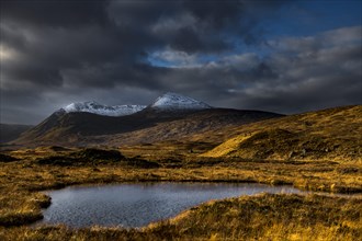 Small lake in the foreground snow-covered mountain peaks of Meall aÂ´Bhuiridh and Clach Leathad in the background and dramatic clouds