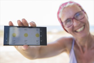 Laughing woman on the beach happy about sunny weather in the weather forecast on her smartphone