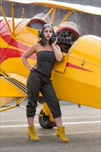 Young woman in green jumpsuit and old flying cap poses with yellow biplane