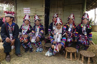 Akha women and men sitting in traditional costumes in front of a bamboo hut