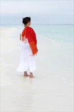 Woman with a red scarf standing on a white beach looking at the turquoise blue sea