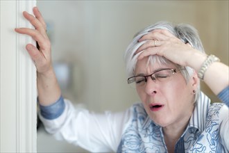 Senior woman with a headache holds her hand on her forehead and props herself on the door frame