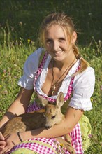 Woman in dirndl with a tame fawn sitting in her lap in a flower meadow