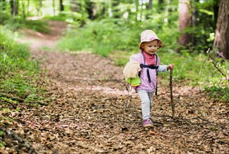 Toddler with doll and walking stick hiking through the woods