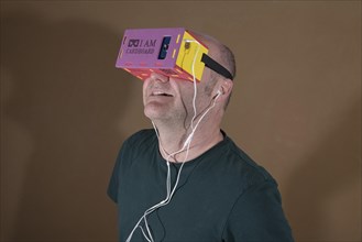Man wearing I AM CARDBOARD 45mm focal length virtual reality Google cardboard VR goggles with Samsung Galaxy S5 Android smartphone and in-ear headphones