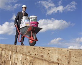Man wearing a suit and helmet pushing a wheelbarrow with building materials along a wall