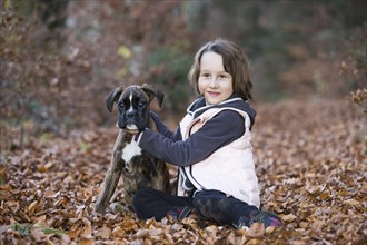 Boxer puppy sitting with little girl in autumn leaves